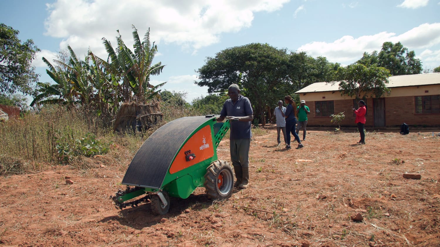 A farmer walking the Aftrak tractor with its solar panel canopy across red soil at Emisizini, houses and trees behind