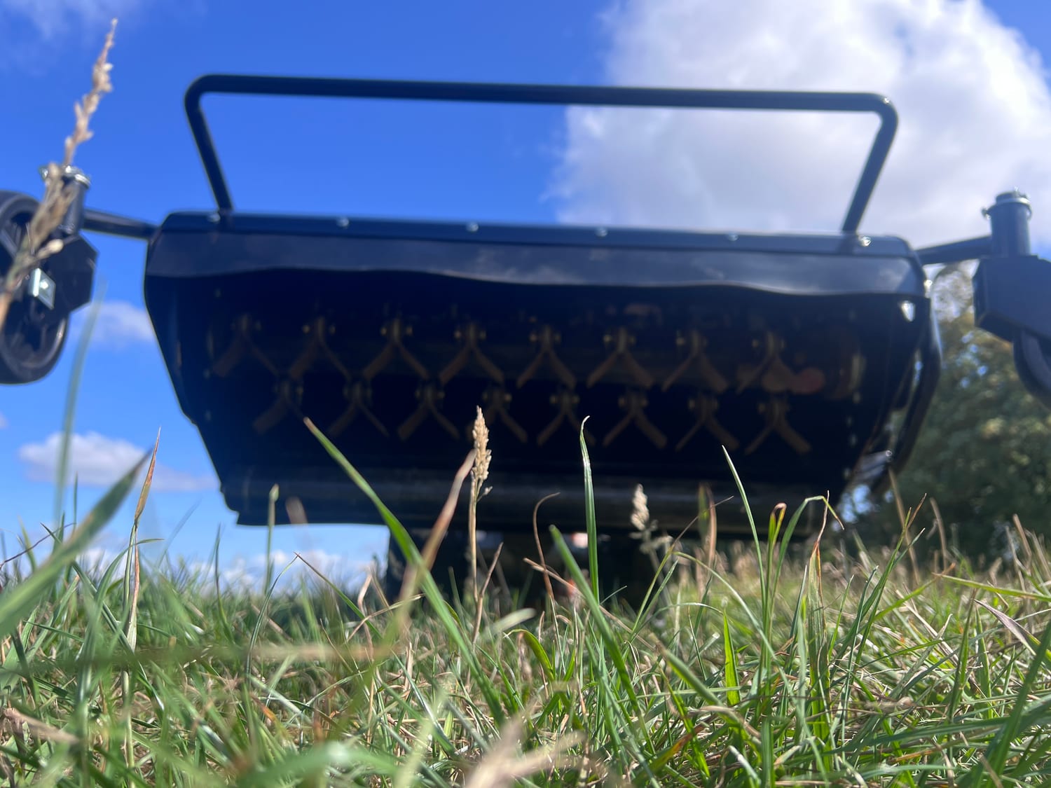 The flail mower from ground level: the black housing with its rows of blades, standing in cut grass under a blue sky