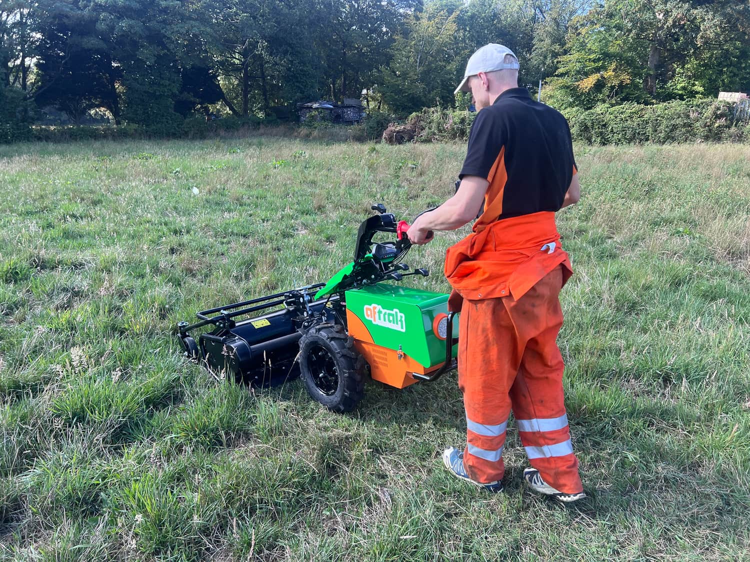 An operator in hi-vis trousers walking the Aftrak tractor and flail mower through long grass at a field edge