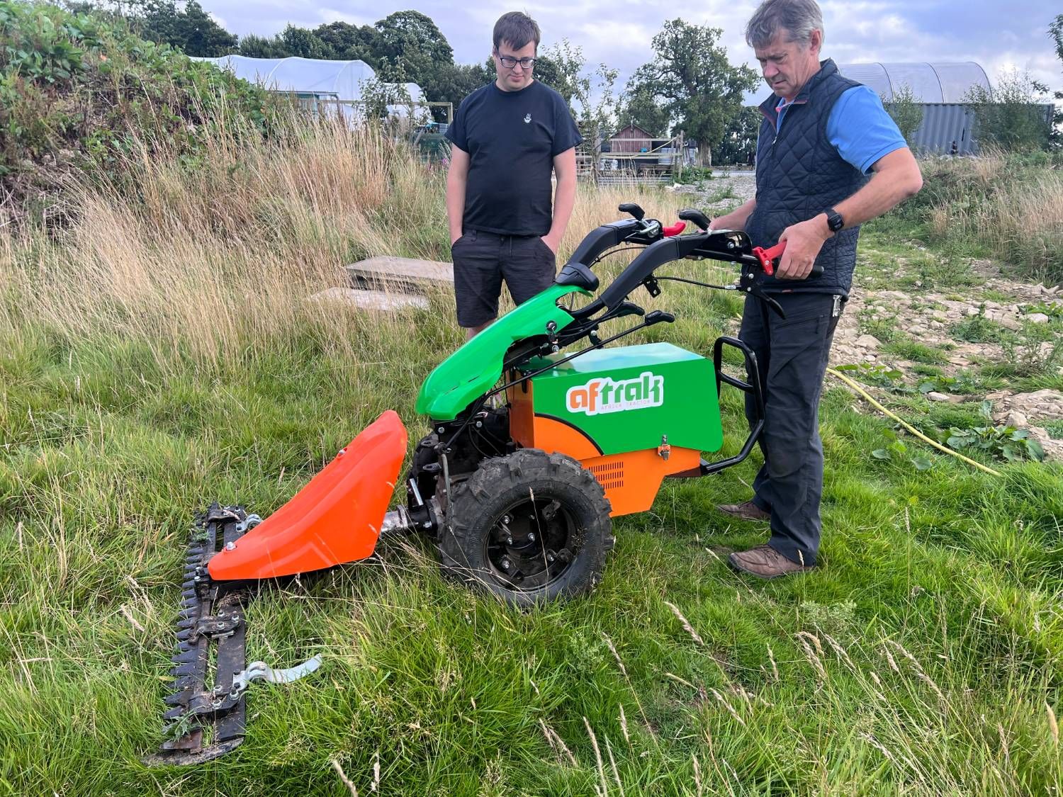 One man at the Aftrak tractor’s handlebars with the cutter bar fitted, another watching, both standing in long uncut grass with the nursery polytunnels in the background