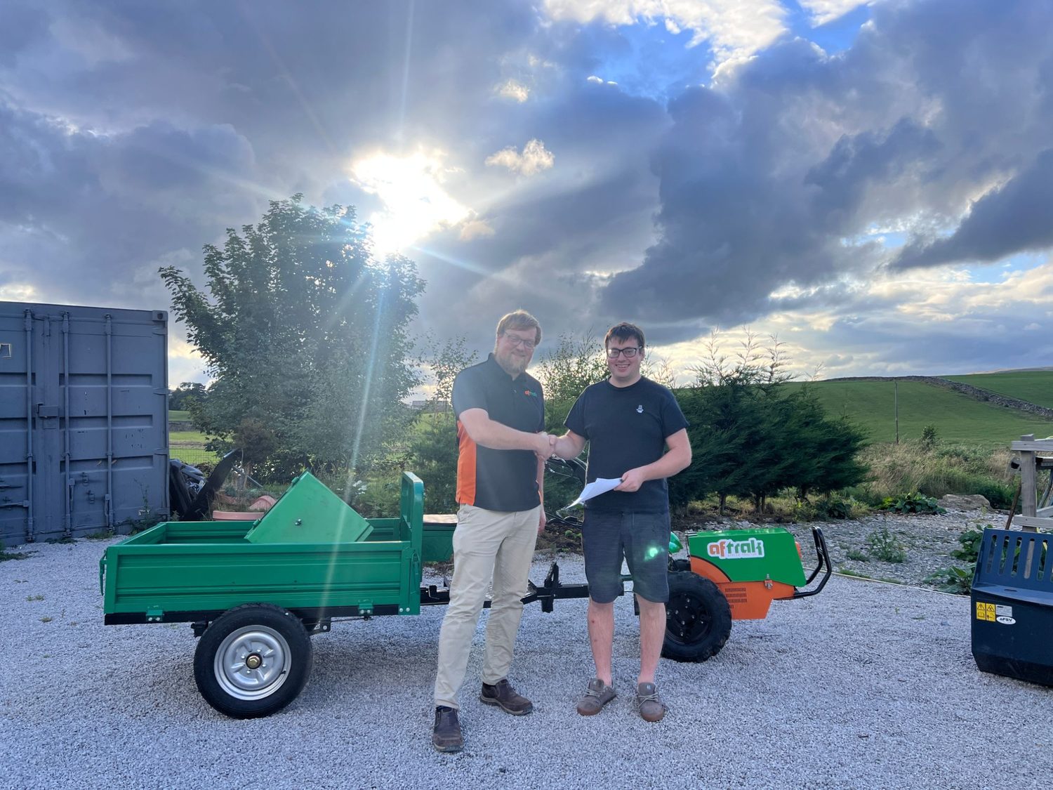 Two men shaking hands on gravel beside the Aftrak tractor and its green tipping trailer, handover paperwork in one hand, low sun breaking through cloud behind them
