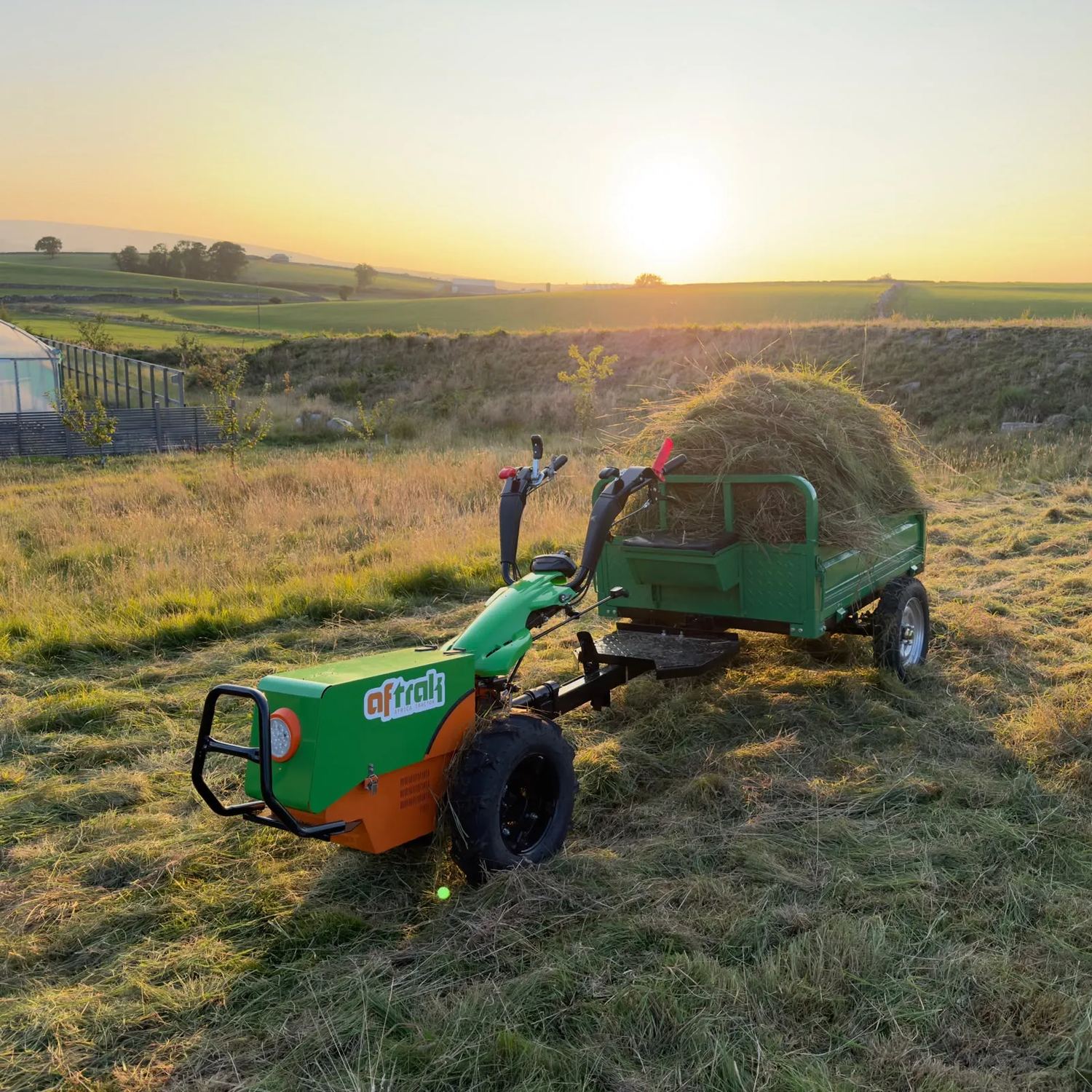 Aftrak tractor and trailer loaded high with cut grass, standing in a mown meadow at sunset with the Dales behind