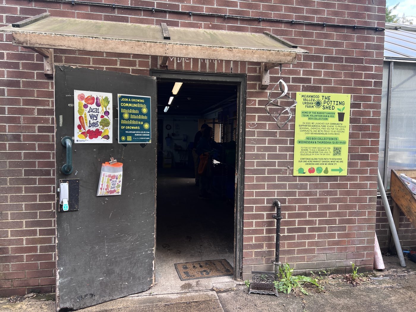 Open door of a brick outbuilding signed The Potting Shed, veg box collection notices beside it