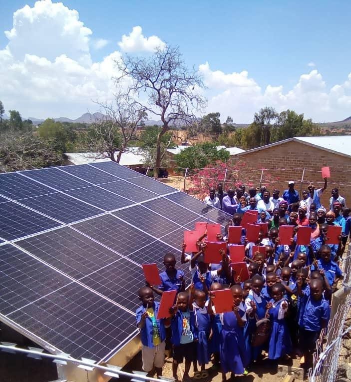 Pupils in blue uniforms gathered beside a large ground-mounted solar array in the school yard