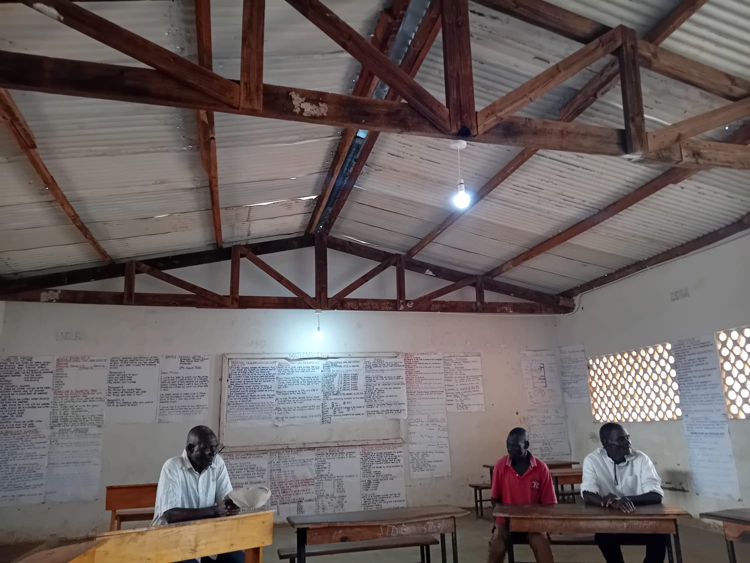 Inside a classroom at Ehlekweni school with an electric light on, three men at desks under a corrugated roof