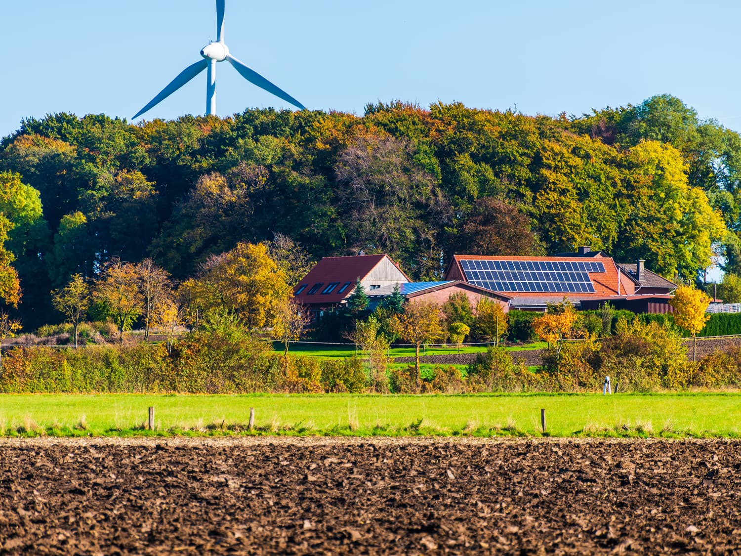 A farmhouse and barn with solar panels on the roof, a wind turbine behind and a ploughed field in front