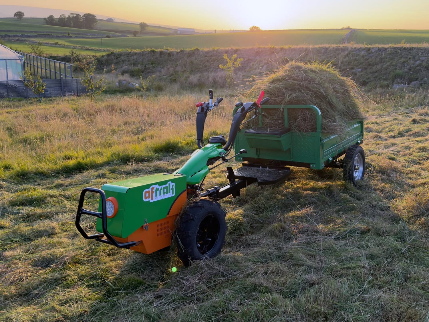 The Aftrak tractor and its green trailer heaped with cut grass in a meadow at sunset