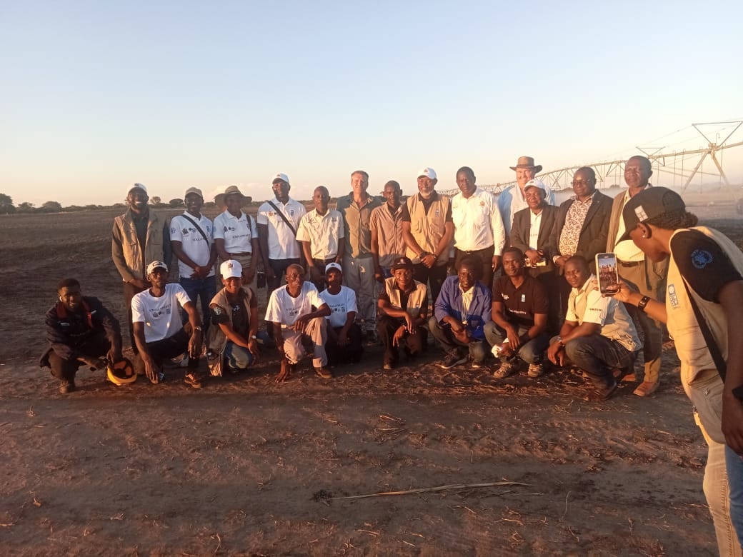 The project team gathered for a group photograph on bare irrigated ground in western Zambia at dusk, a centre-pivot irrigator behind them